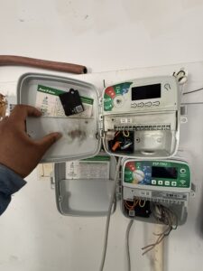 Inspecting the exposed wiring compartment of a wall-mounted Rain Bird sprinkler controller that was damaged by a lightning strike.