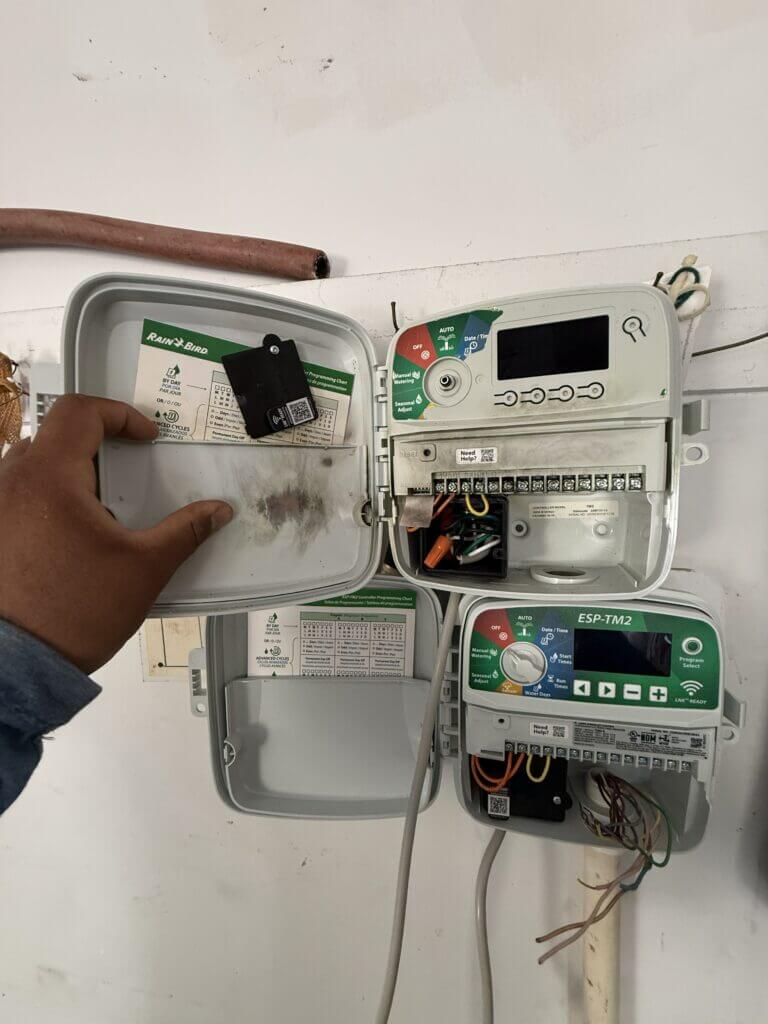 Inspecting the exposed wiring compartment of a wall-mounted Rain Bird sprinkler controller that was damaged by a lightning strike.