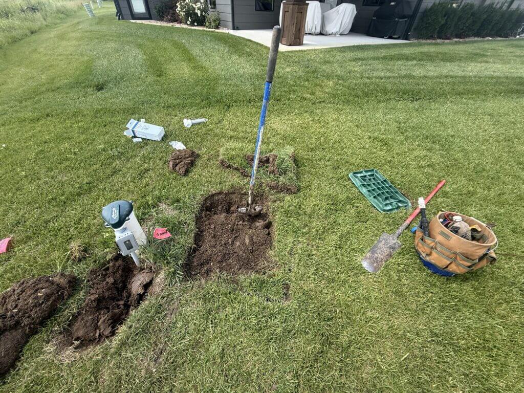 Exterior view of a green lawn with a newly dug hole for an irrigation system repair, featuring scattered shovels, a tool bag, and flags.