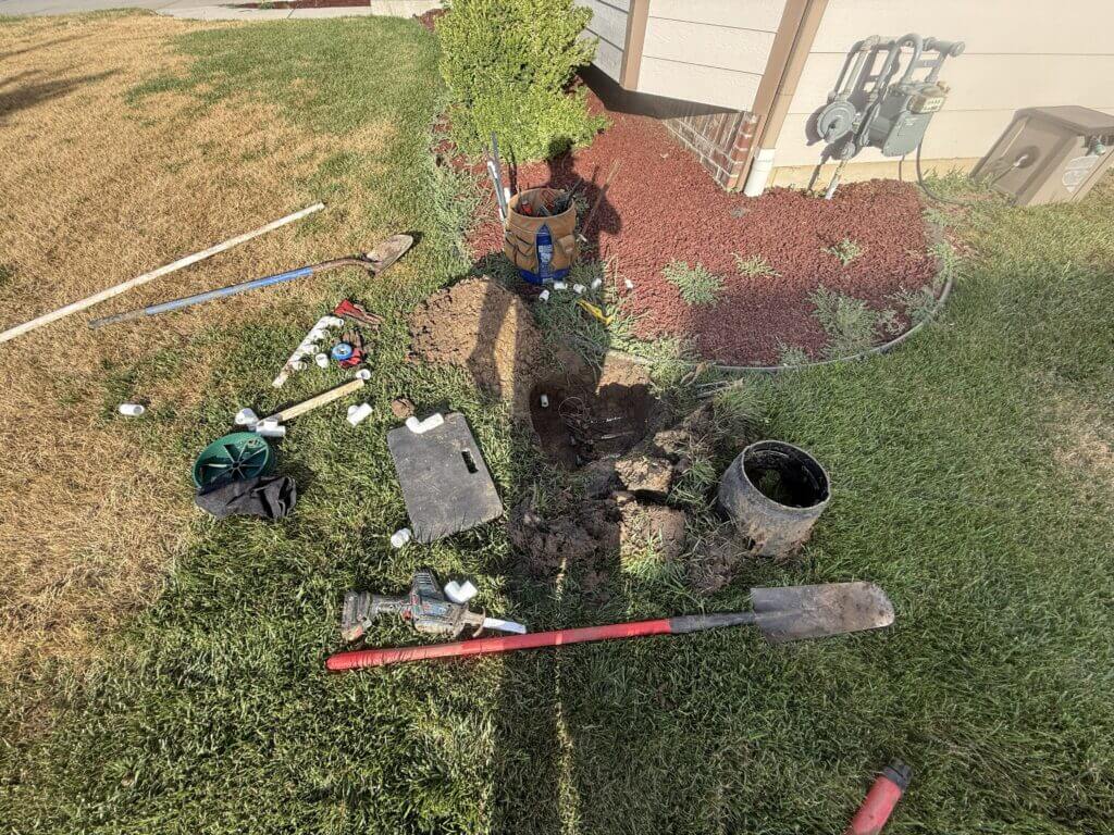 A landscape repair site near a house corner, with red mulch, a tool bucket, and scattered tools like a shovel and saw on the grass near an excavated hole.