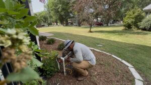 An irrigation technician crouching next to a house foundation, using a test gauge and hoses to perform a backflow test on a lawn sprinkler system.