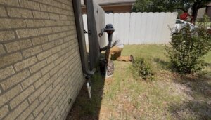 An irrigation technician crouching near a wooden privacy fence to perform a residential backflow test on a sprinkler system where he is about to turn on the water.